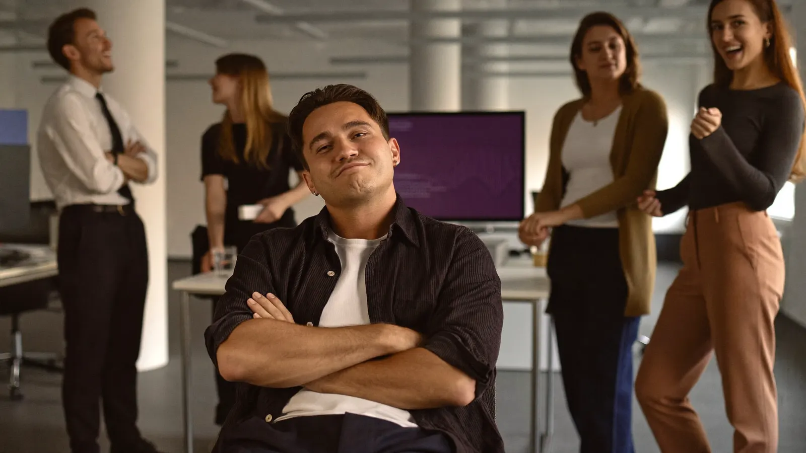 A confident coworker with arms crossed while colleagues laugh behind him